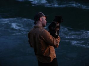 Country singer Zach Bryan stands in front of the ocean holding a dog