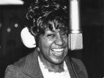 Image of POC songwriter Rose Marie McCoy smiling behind a microphone