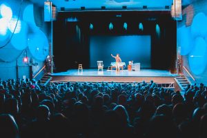 Image of a person on stage and a crowd watching at a theater