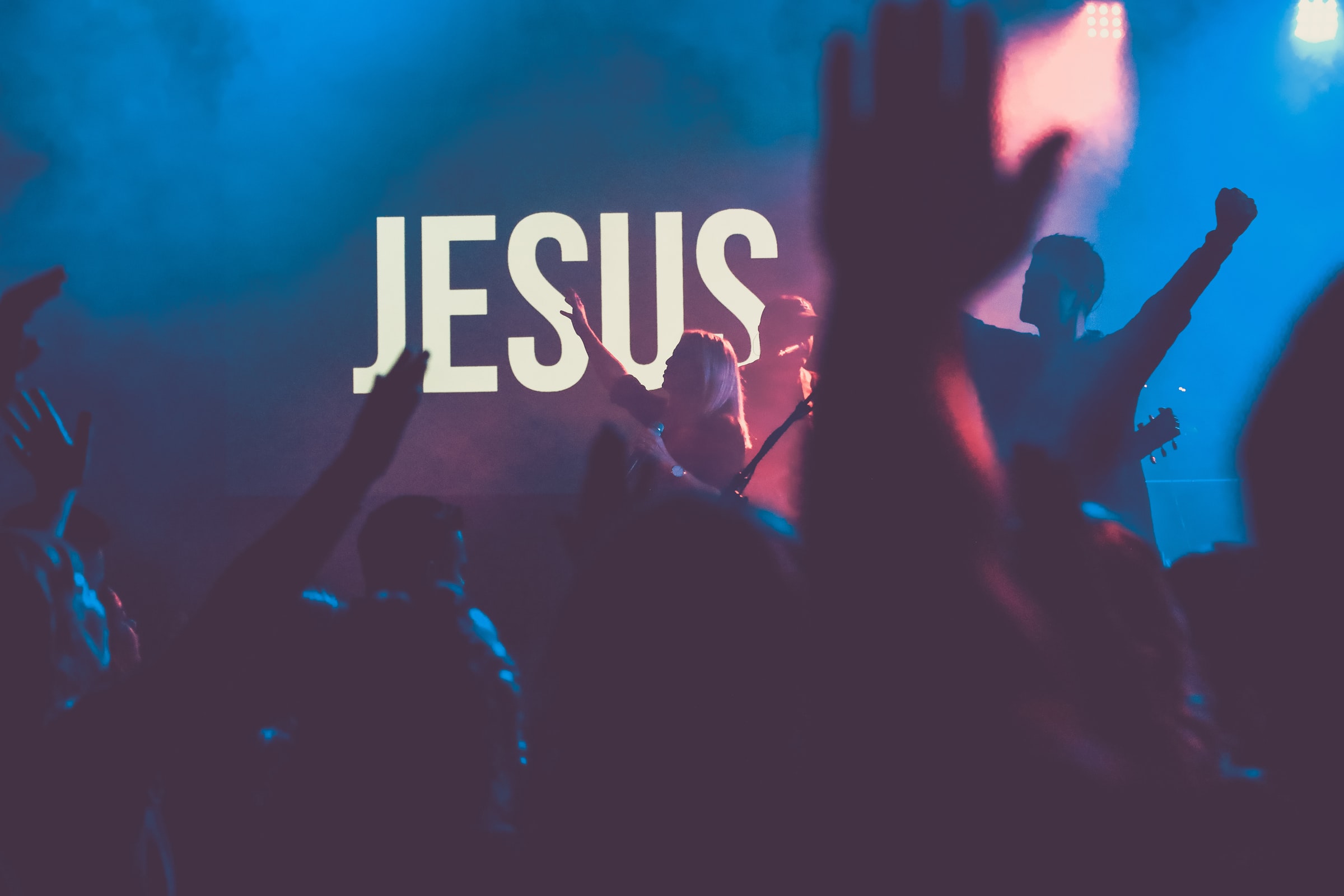 An image of a megachurch service, depicting a crowd of people raising their hands in worship before a sign reading "Jesus" and a Christian rock band performing onstage.