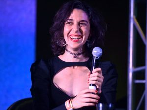 In an article about Jamie Loftus, the comedian stands against a blue and black background while holding a microphone.