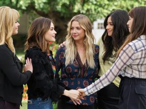 In an article about Pretty Little Liars, five woman stand holding hands and touching shoulders in a garden.