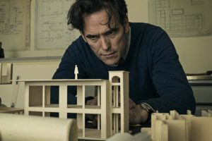 A screenshot from The House That Jack Built in which serial killer Jack looks over a model of the house he plans to build throughout the film.