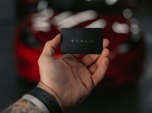 In an article about Elon Musk, a hand holds a black Tesla card in front of a blurry red Tesla.