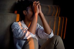 A person is sitting and leaning against a wall with their hands clasped to their forehead in a stressed-out position.