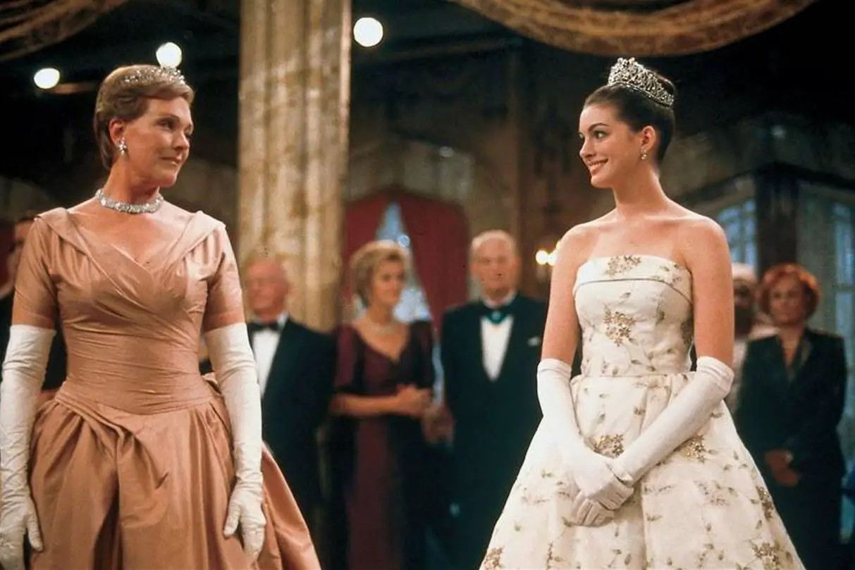 In an article about Anne Hathaway's Fairy-Tale Films, a screenshot of Anne Hathaway in the Princess Diaries dressed in royal clothing at a ball next to Julie Andrews