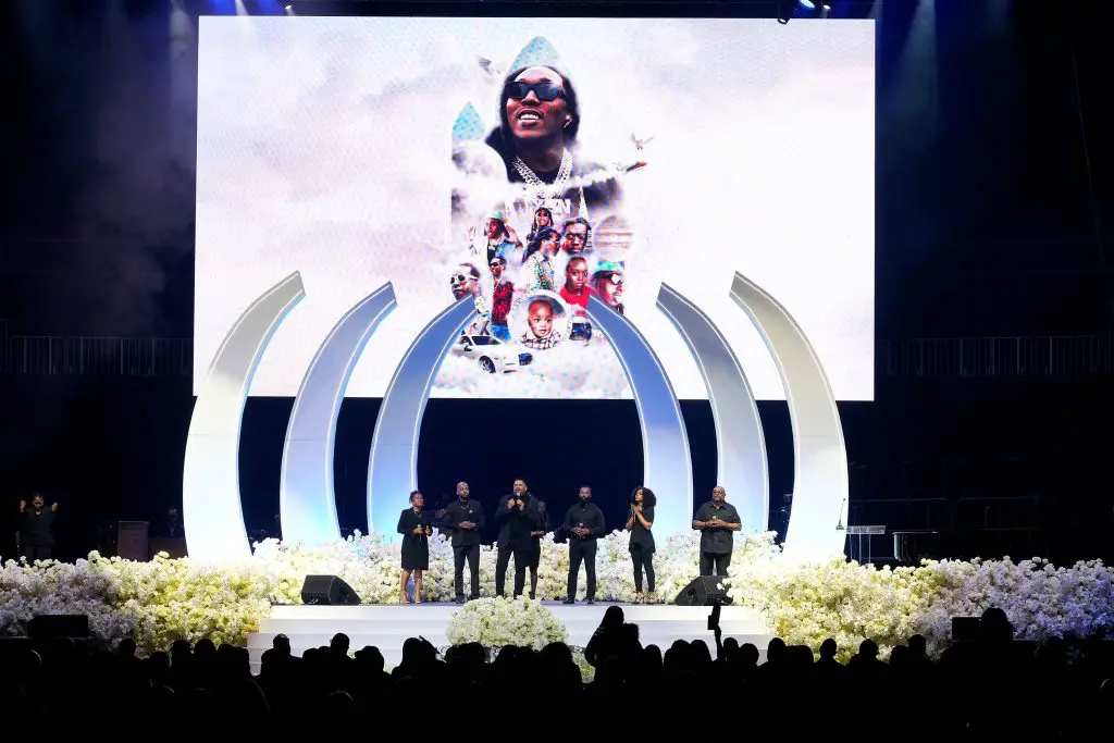 A screenshot from takeoff's funeral shows a group standing on stage in front of his picture on a screen.