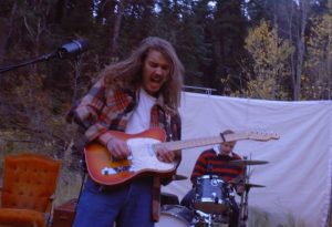 In an article about The Backseat Lovers, the band's guitarist plays in front of what appears to be a white sheet. A drummer can be seen in the background.