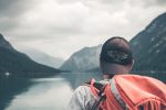 In an article about travel YouTubers, a man with an orange backpack looks out on a lake.