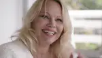 In an article about "Pamela, a love story" is a screenshot from the documentary that shows Pamela Anderson in a white shirt and behind a green garden, smiling.