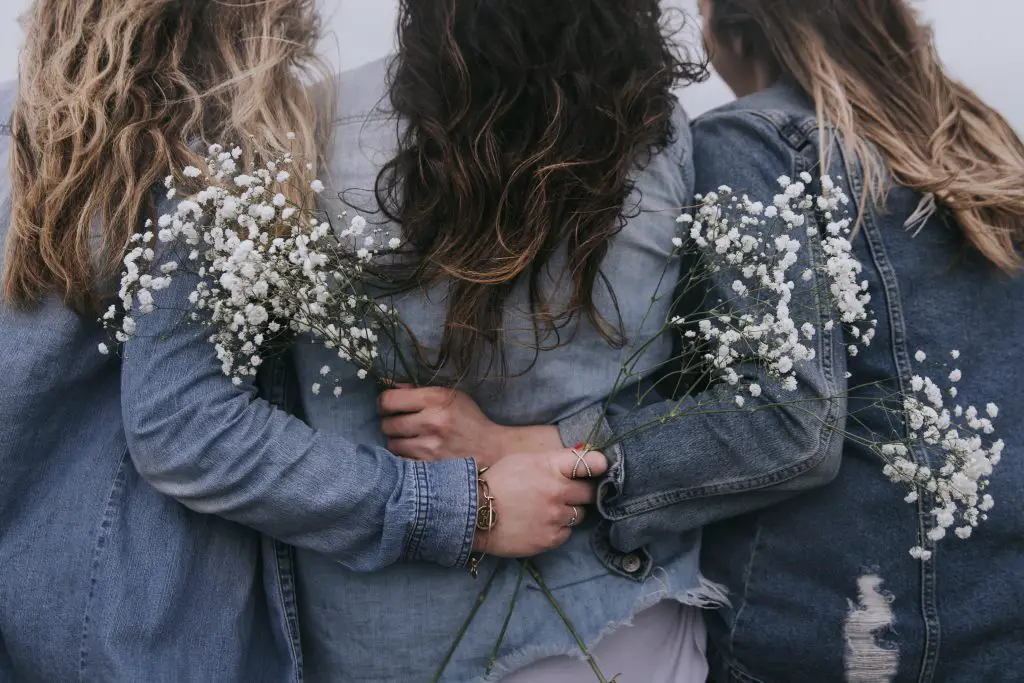 In an article on Women's History Month is a photograph of three women in jean jackets facing backward and holding white flowers.