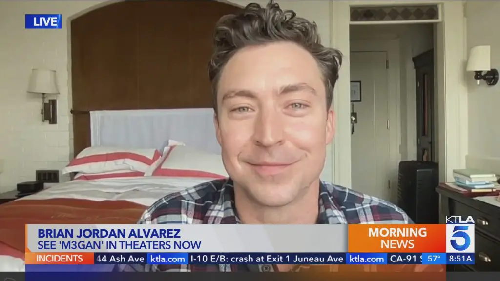 A close-up of actor Brian Jordan Alvarez smiling from inside a bedroom. Below his face is a grey, orange, and blue news banner with the words "Brian Jordan Alvarez" and "See 'M3GAN' in theaters now" in blue font.