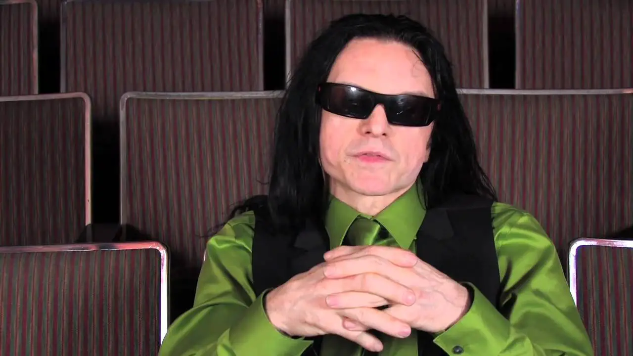 In an article about "The Room," a white man with long black hair, wearing dark sunglasses and a bright green button down shirt, sits in an auditorium with his hands clasped in front of him.