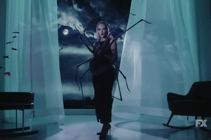 In an article about Kim Kardashian in "American Horror Story Delicate," Kim Kardashian stands in a black dress with a white wig on in front of draping white curtains in blue-ish lighting.