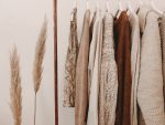 In an article about thrift stores, a clothes rack with sweaters in various shades of beige hanging from it. To the left of the clothes rack are two tan pieces of wheat.
