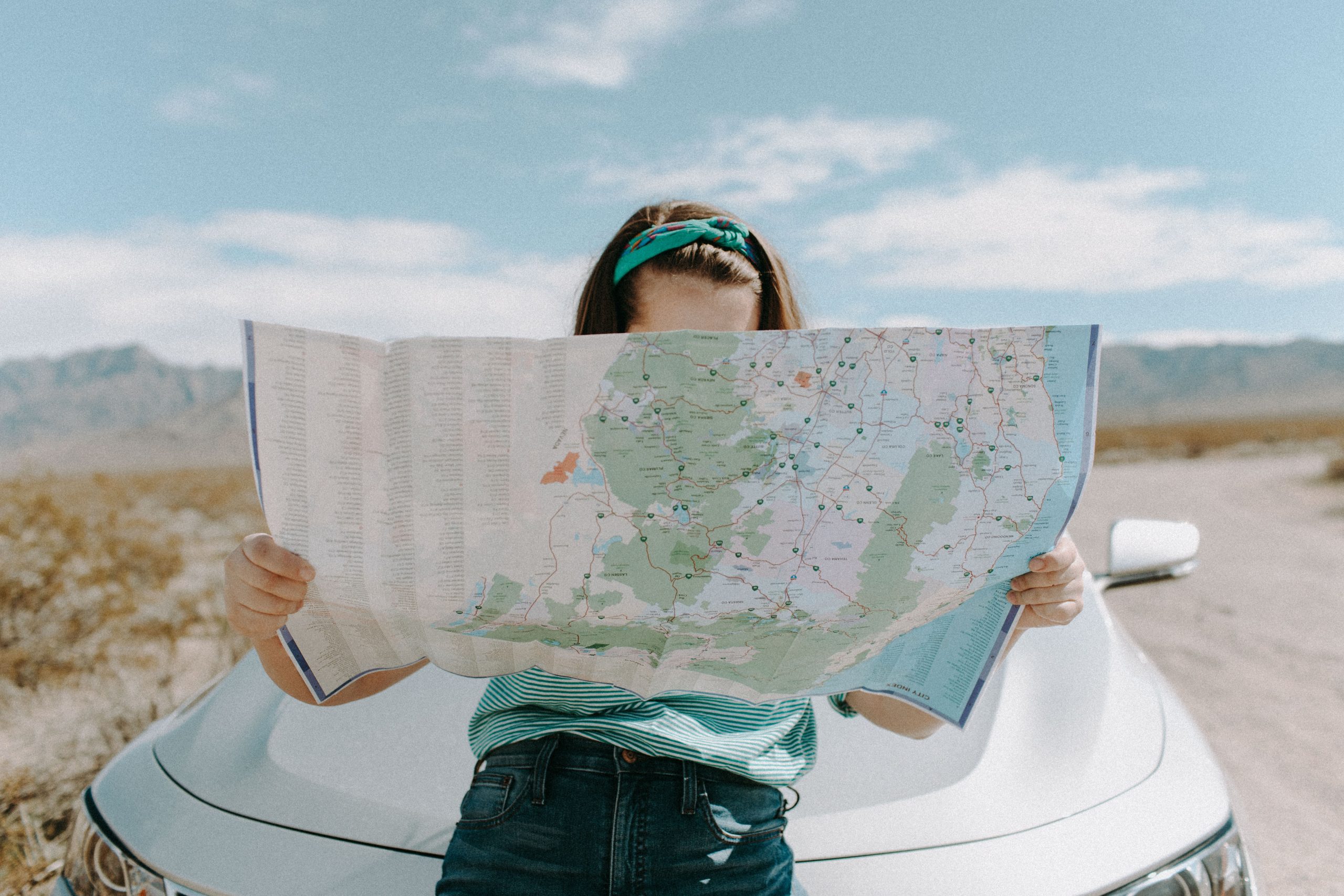 In an article that gives tips on traveling the world as a broke college student is a photograph a woman holding a roadmap and standing outside of her white car on a desert road.