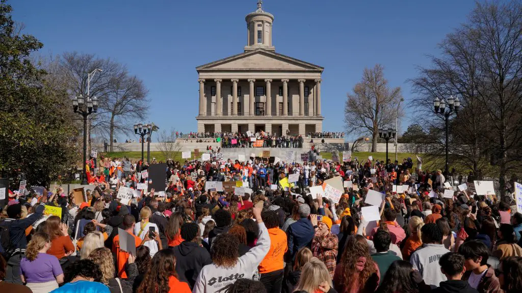 In an article about the Nashville school shooting, a crowd of protesters holding up signs face a large, stately-looking tan building with many large columns and carved details.