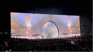Beyonce is reflected in a bright double image across a giant screen, in front of an audience of hundreds in a darkened stadium.