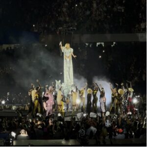Wearing a long, glittering silver dress, Beyonce stands on a raised platform above her backup dancers and fans.