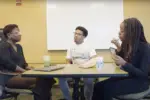 In an Article about web series, "Reality Check," three people sit at a desk. One applies makeup sitting across from one who speaks as another looks on.