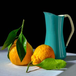 In an article about paintings, two oranges and a blue pitcher sit on a table against a black background.