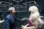 In an article about Mattel's use of self deprecation, a still from the 'Barbie' movie. Will Ferrel portrays Mattel CEO and Margot Robbie portrays Barbie. He Stands in a black suit at the center with a pink tie. He carries a letter opener in his hand. They stand in a labyrinth, screaming in each others face.