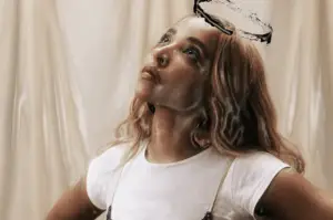 In an article about Tinashe's new album 'BB/Angel,' a woman with blonde hair looks up. She wears a white tee shirt with a slip dress over it. A hand-drawn angel halo is above her head.