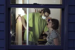 In an article about 24-hour homecare worker shifts, a homecare worker wears a lime green shirt and a face mask while helping a woman in glasses and a black and white striped shirt who sits on a bed.