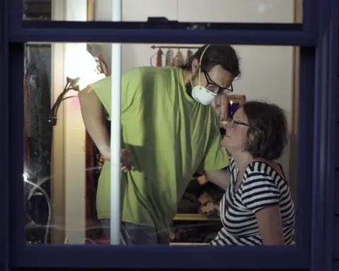 In an article about 24-hour homecare worker shifts, a homecare worker wears a lime green shirt and a face mask while helping a woman in glasses and a black and white striped shirt who sits on a bed.