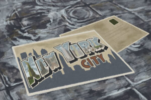 In an article about two essays about New York City, a post card with the words "New York City" across a cityscape. The post card is in a grey rain puddle.