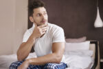 Man sitting on edge of bed with hand on chin thinking.