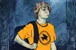 For an article about the Percy Jackson TV series, a boy with mid-length brown hair wears a bright orange-yellow shirt with a pegasus (winged horse) on it while carrying a dark backpack. He stands against a blue, water-like, crinkly paper background with a Greek-style border along the left and right side.