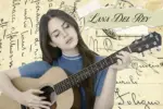 A woman with brown hair and a blue sweater is sitting in front of a background of paper covered in writing, intimately strumming a guitar. Her eyes are closed and her lips set in a stoic position, implying she is in deep contemplative as she parcels out each individual note. Her name -- Lana Del Rey -- is written on a torn piece of paper, taped to the background in the upper-right corner.
