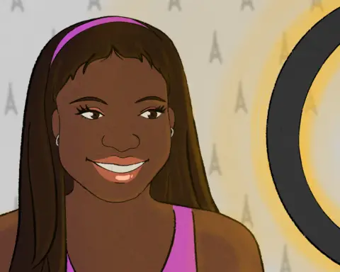 A woman with brown skin, dark hair and a pink headband stands in front of a grey wall, with A-shaped markings in it. She's smiling into a camera stand from which the silhouette of an iPhone can be seen; the ring of the stand glows yellow, presumably casting light on her as the phone's camera captures her in shot.