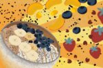 A white bowl of food dusted with grey sits in front of an orange-and-yellow striped background. It contains substances of various colors: white, brown, clay-esque in tone, a caramel stripe running through the center. Bananas and blueberries top it, whilst other fruits such as strawberries, more blueberries and peaches explode across the back ground, black flecks flying with them.